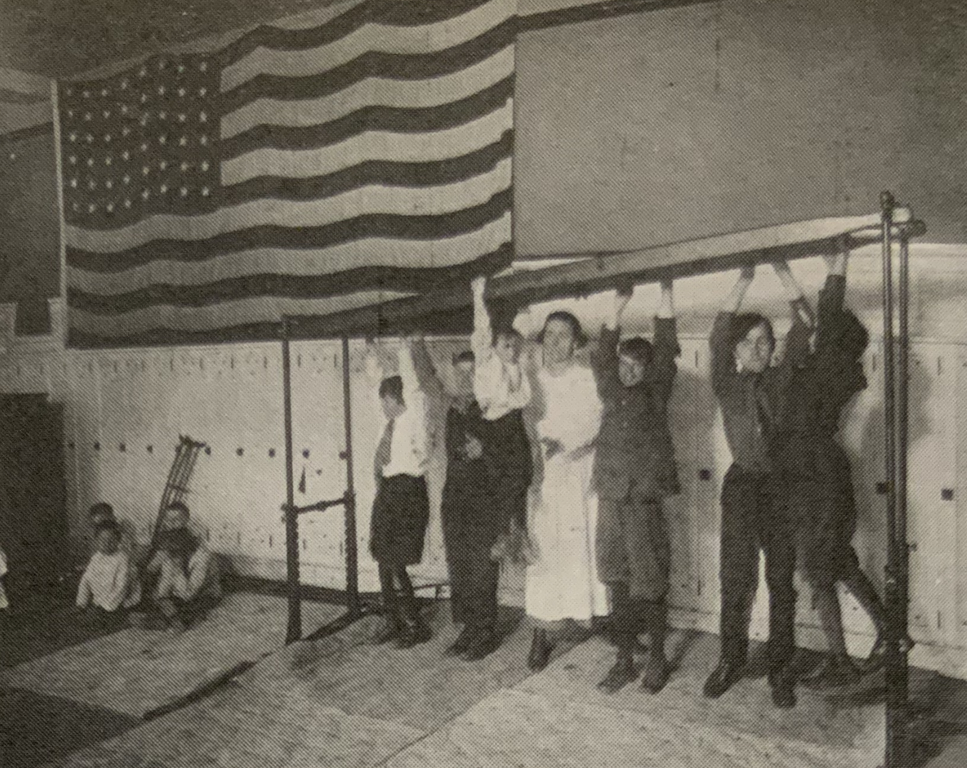 students hand from a gymnatsics bar during Physical therapy in the gym. there is a a large american flag on the wall behind them